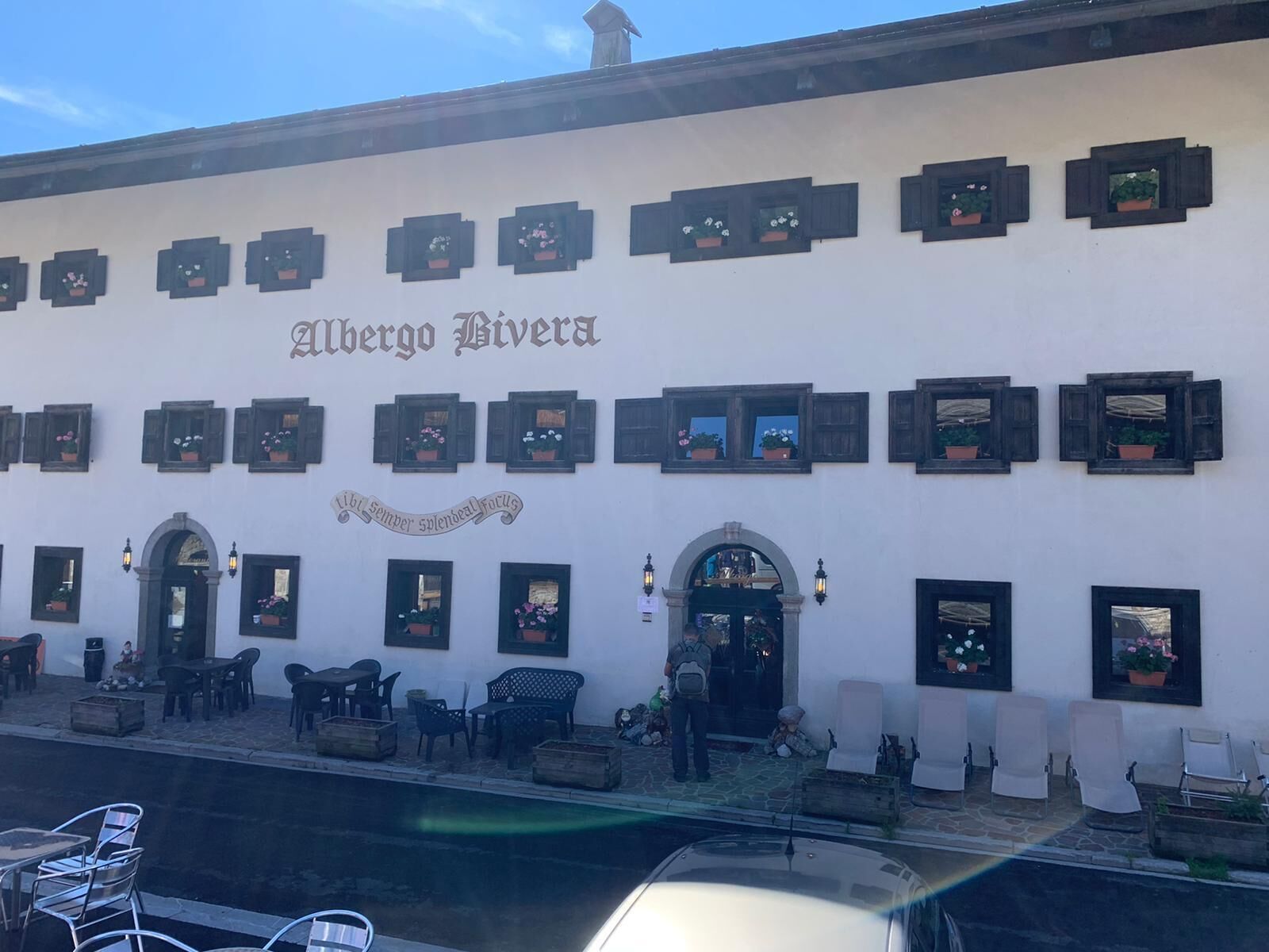 A building with the name "Alberto Riviera" on the facade, featuring multiple windows with flower boxes, outdoor seating, and a stone-paved entrance area.