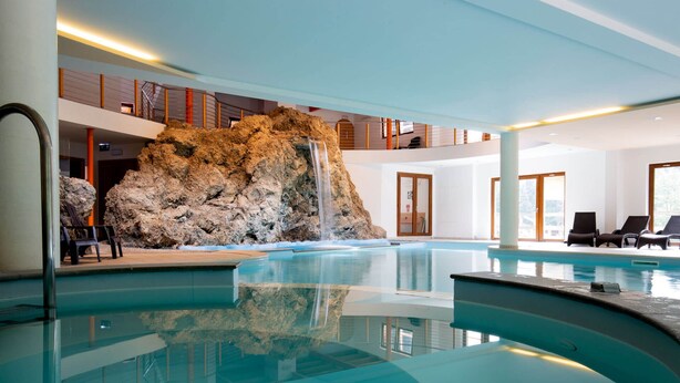 An indoor swimming pool with a waterfall feature, lounge chairs, and a view of the outdoors. - Hotel Bivera Sauris (Sauris)