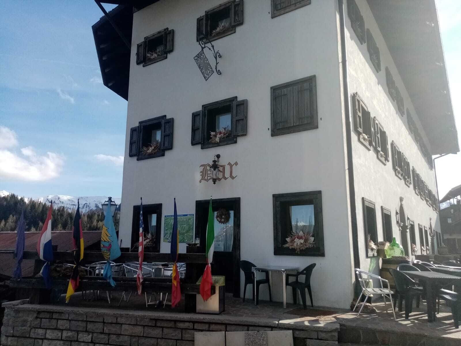 Un edificio con varias ventanas, algunas con contraventanas, y un cartel con un escudo. Hay banderas frente al edificio, y un área de estar al aire libre con mesas y sillas.