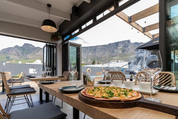 Comedor con una pizza sobre una tabla de madera, mesas, sillas y vistas a montañas a través de amplios ventanales.