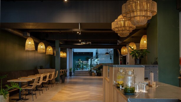 Een restaurantinterieur met houten tafels en stoelen, hangende rieten lampen en een toonbank met een glazen vitrine.