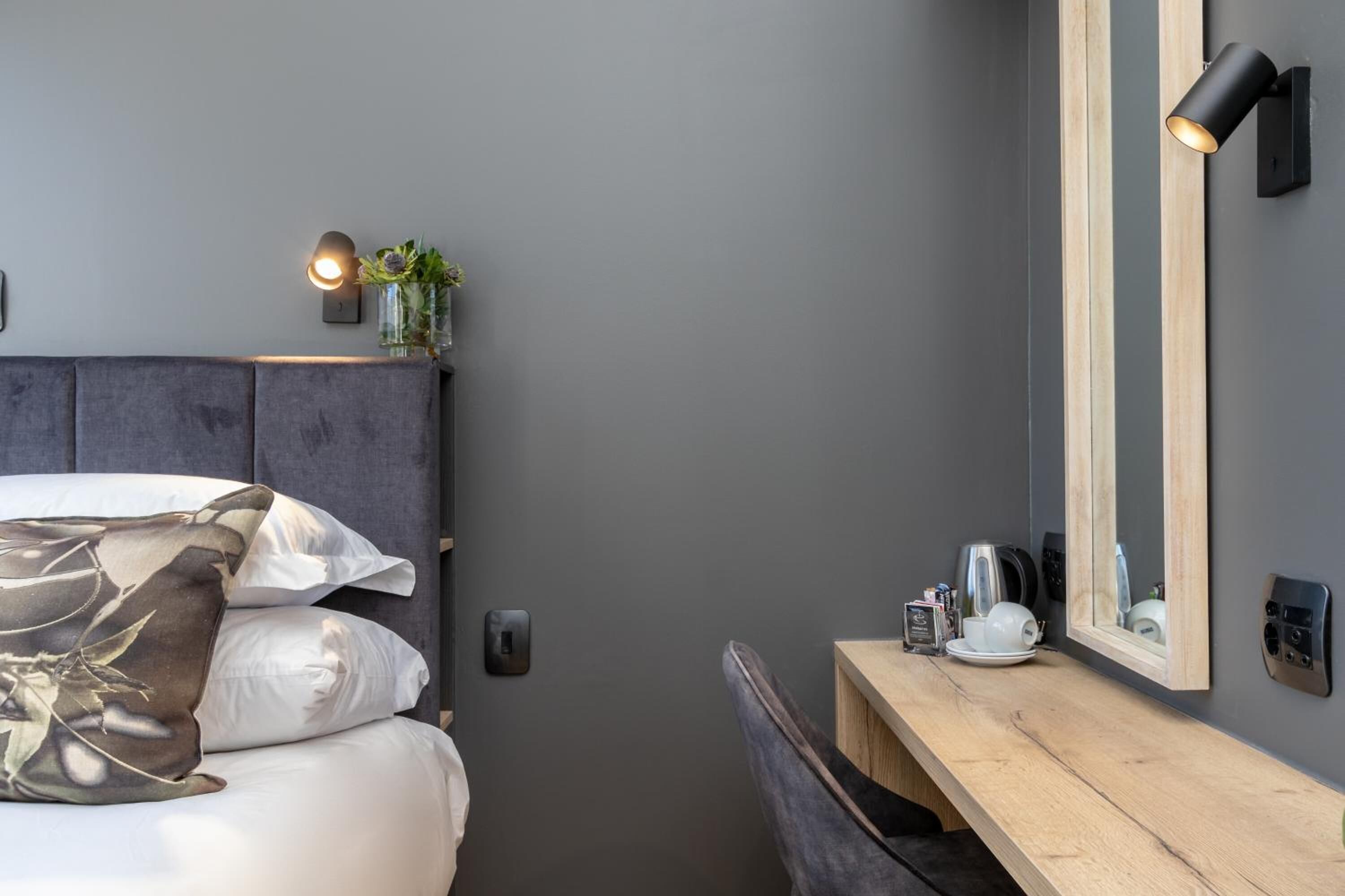 A modern bedroom with a grey headboard, a bedside table with a mirror, and a small plant.
