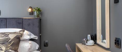 A modern bedroom with a grey headboard, a bedside table with a mirror, and a small plant.