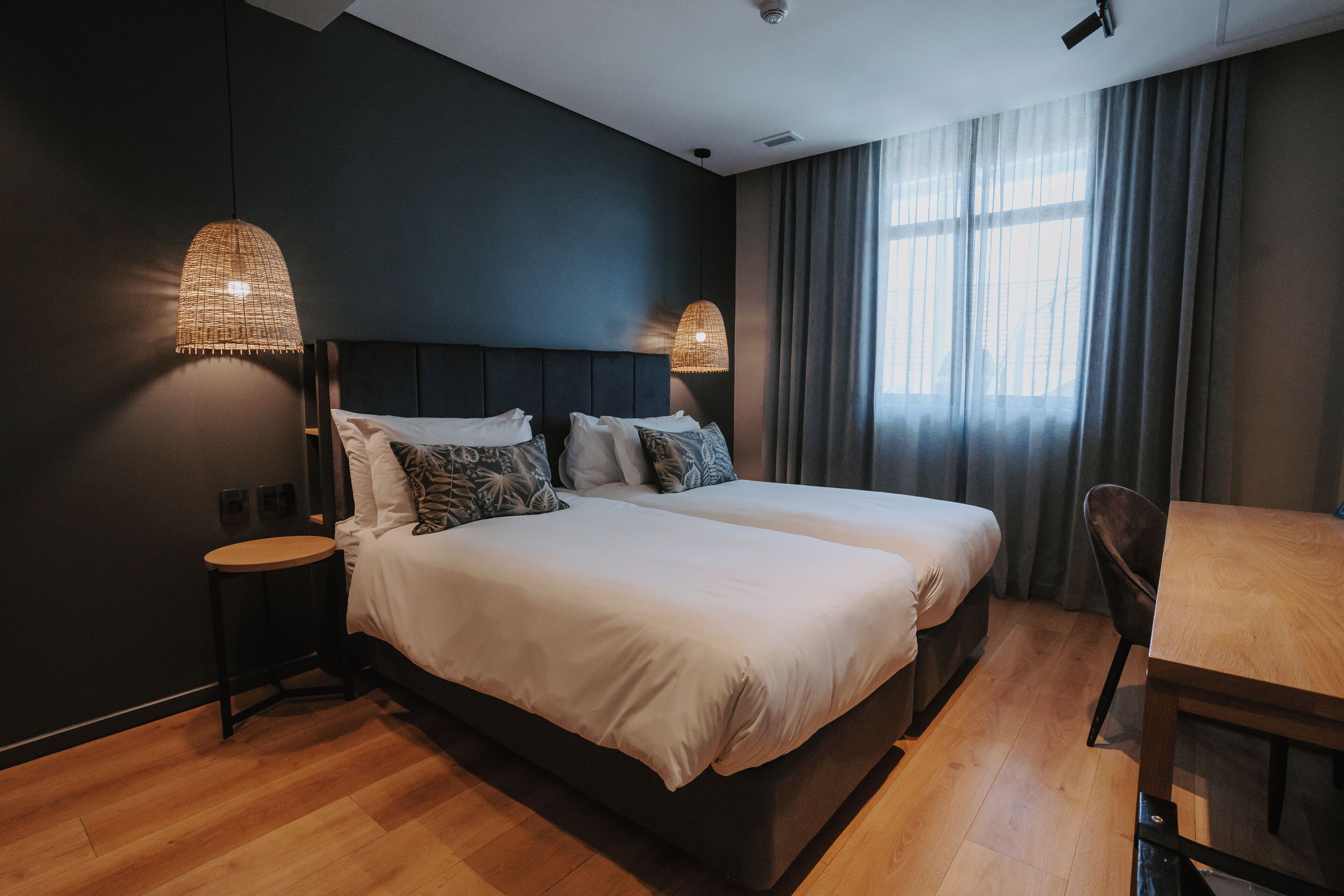 A modern hotel room with a large bed, two bedside tables, a wooden dining table, and a large window with curtains.