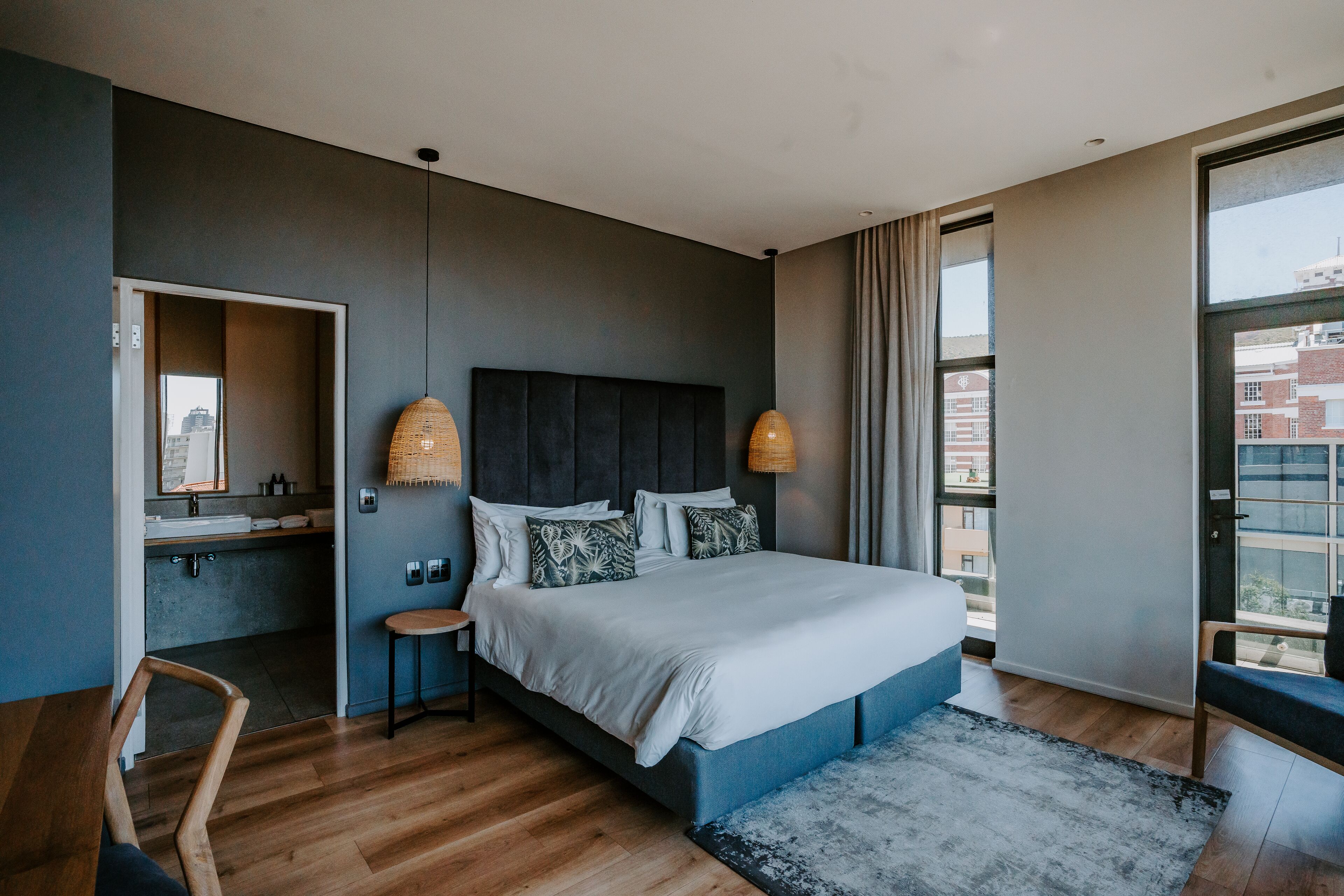 Ein modernes Schlafzimmer mit einem großen Bett, einem Nachttisch, einem Sessel und Blick auf die Stadt durch große Fenster.