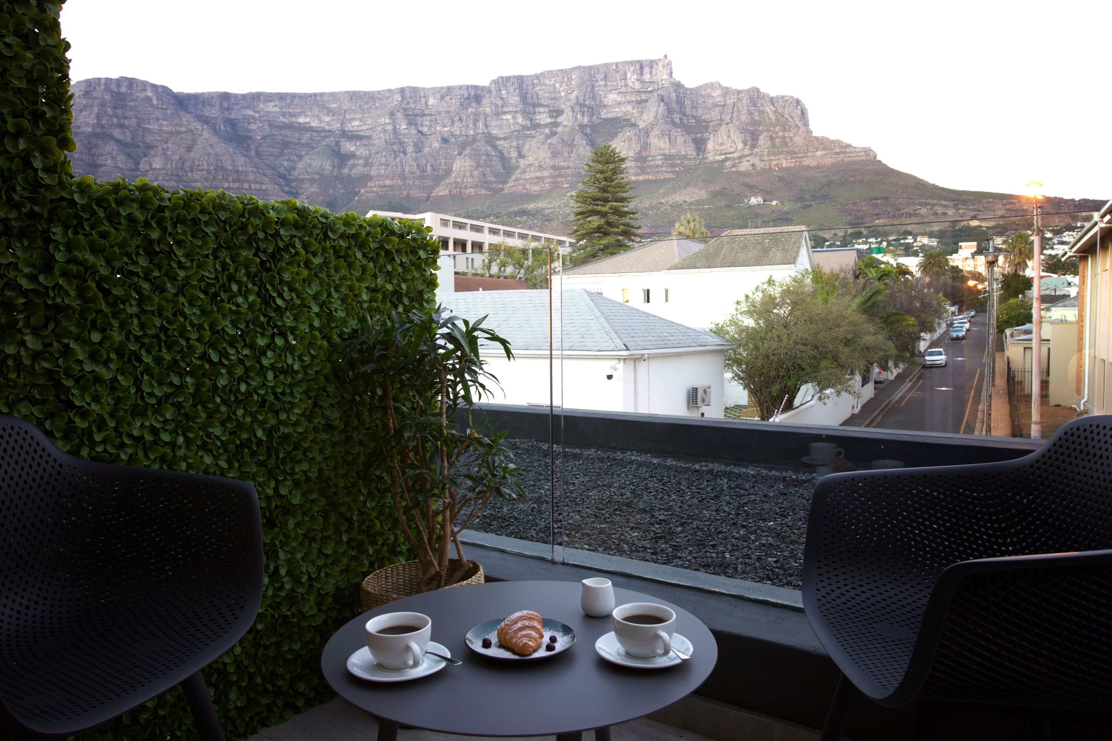 Een balkon met twee stoelen, een tafel met een croissant en koffiekopjes, en uitzicht op een berg en gebouwen.
