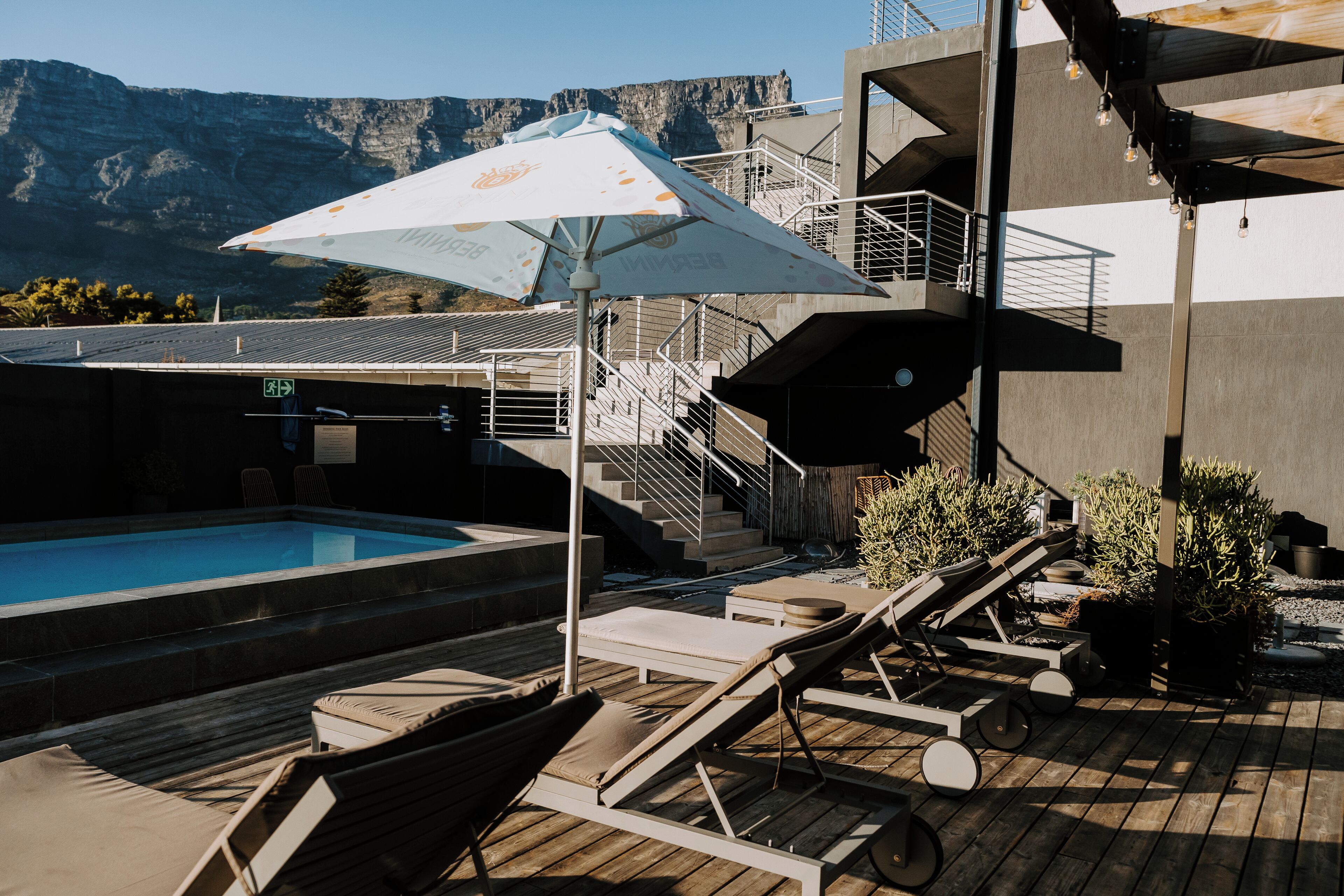 Un espace piscine aménagé avec des chaises longues, un grand parasol et un escalier menant à un niveau supérieur.