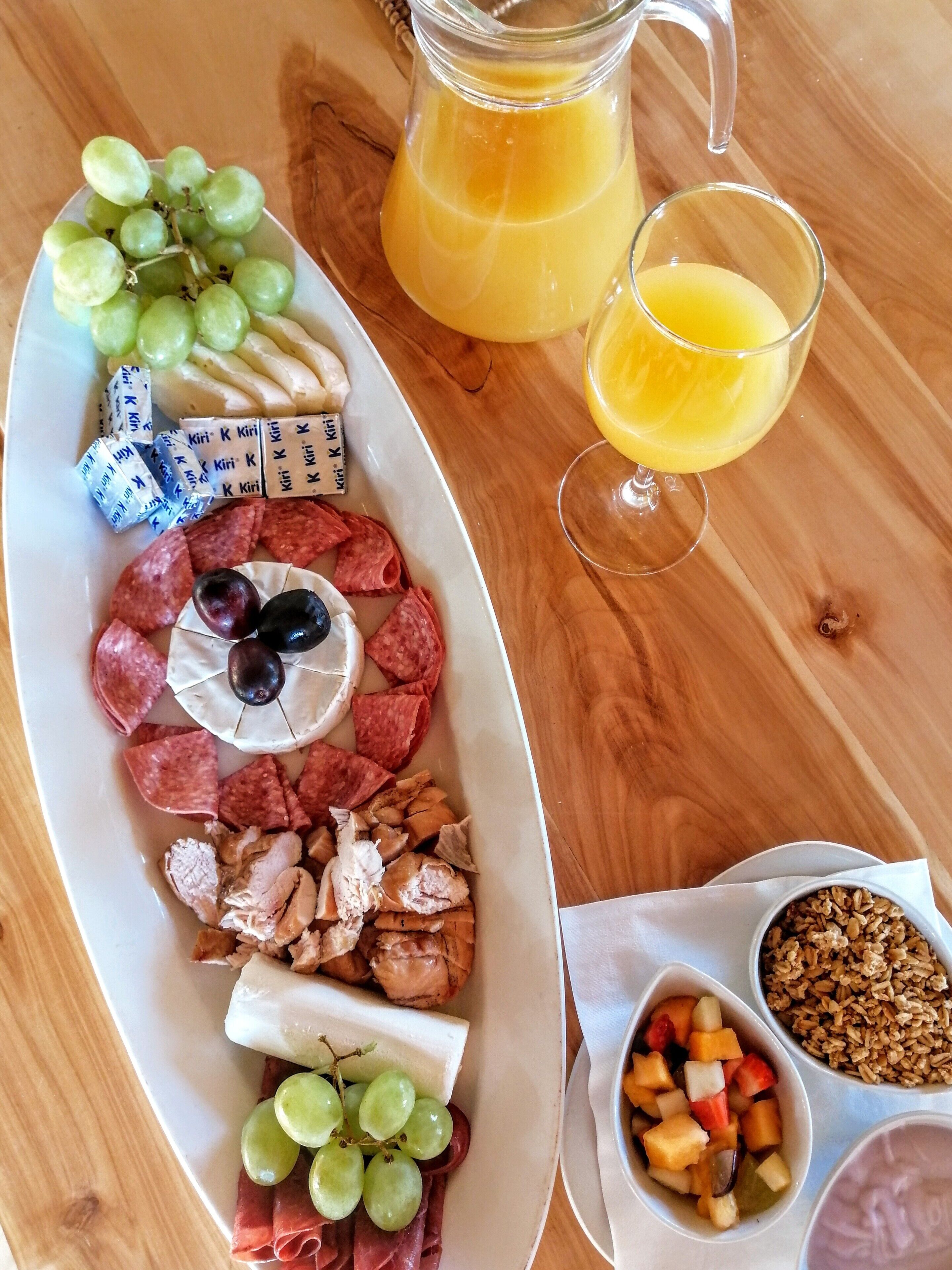 Uma mesa de madeira com uma tábua de frios, um jarro de suco de laranja e tigelas com frutas e granola.