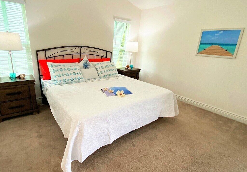 A neatly made bed with a white bedspread and multiple pillows, two bedside tables with lamps, a wooden nightstand, and a framed picture of a beach on the wall.