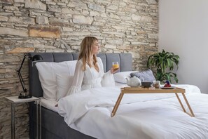 A person sitting on a bed with a tray of breakfast items, a lamp, and a potted plant in a room with a stone wall.