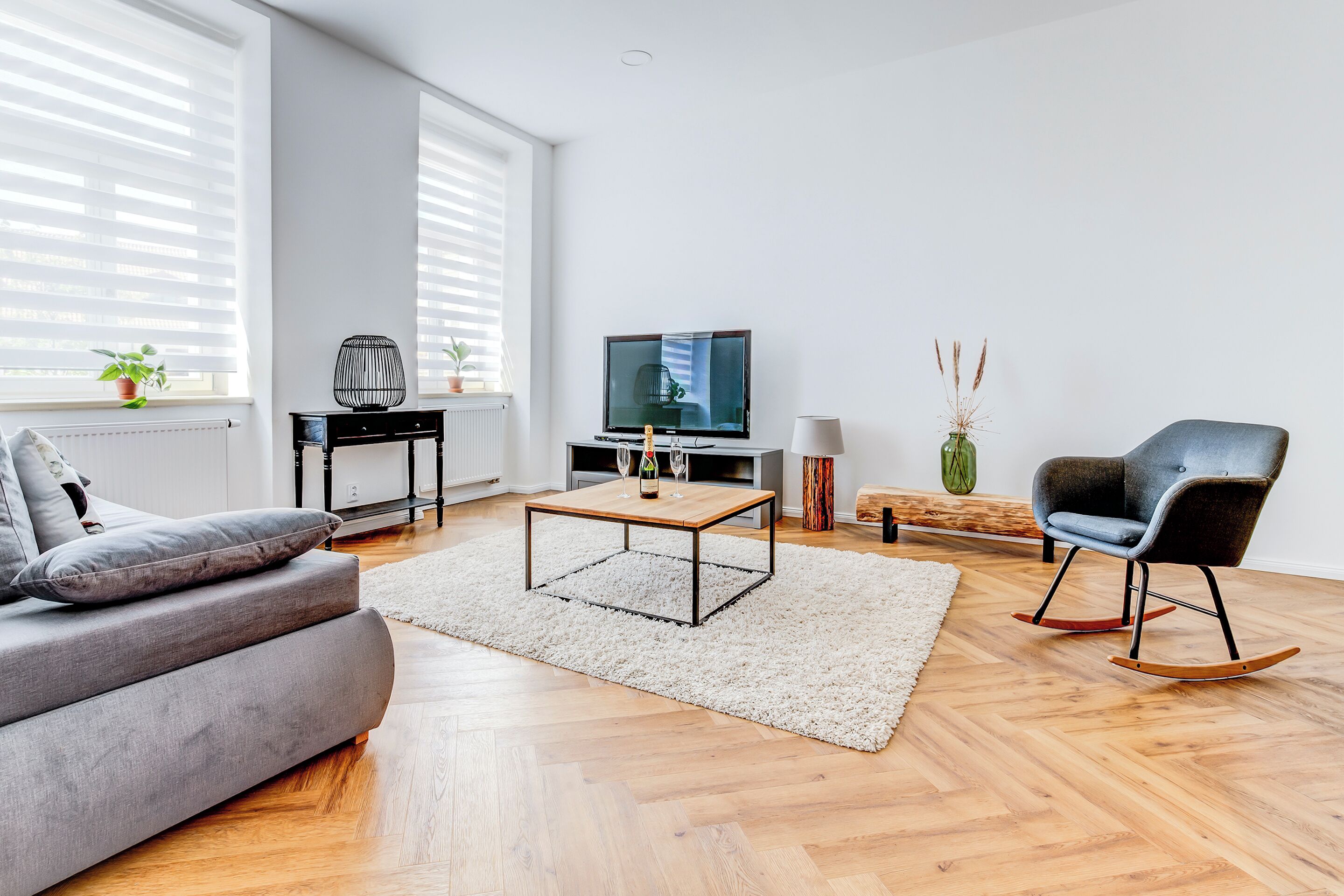 A modern living room with a grey sofa, a wooden coffee table, a TV, and a rocking chair.