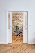 Quarto com piso de madeira em padrão herringbone, uma porta branca que dá para uma parede de pedra e uma planta em cesto trançado.