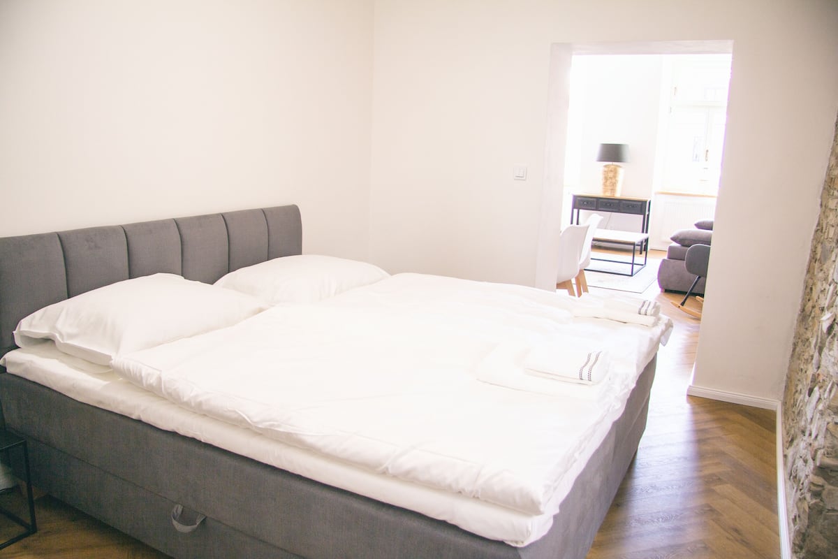 A neatly made bed with a grey headboard, a small bedside table, and a chair in a bright room with a wooden floor.