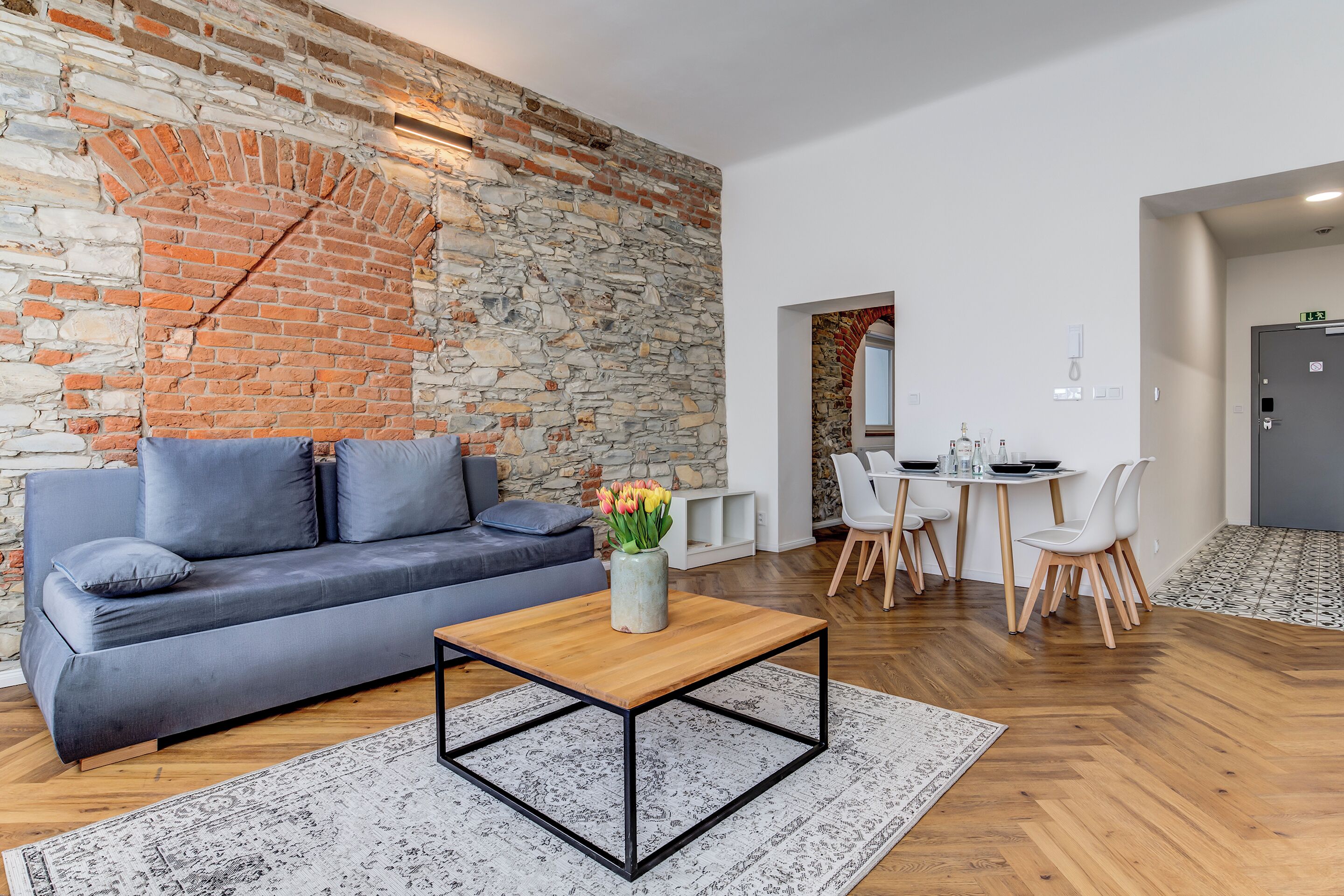 Una sala moderna con pared de piedra, un sofá gris, una mesa de centro de madera y un comedor con sillas blancas y una mesa.