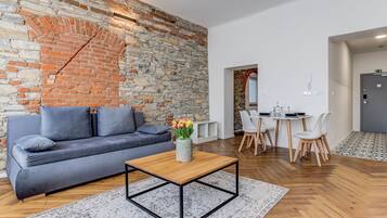 Una sala moderna con pared de piedra, un sofá gris, una mesa de centro de madera y un comedor con sillas blancas y una mesa.