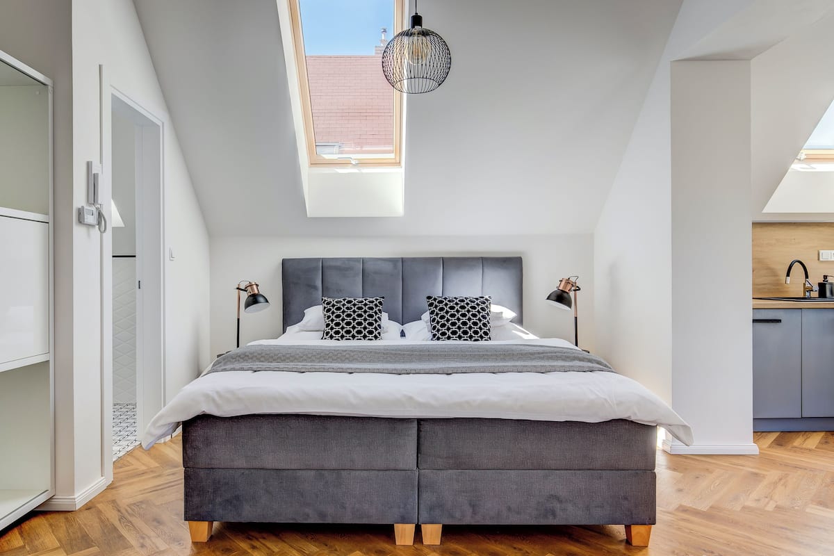 A modern bedroom with a large bed, a skylight, and a view of the kitchen.