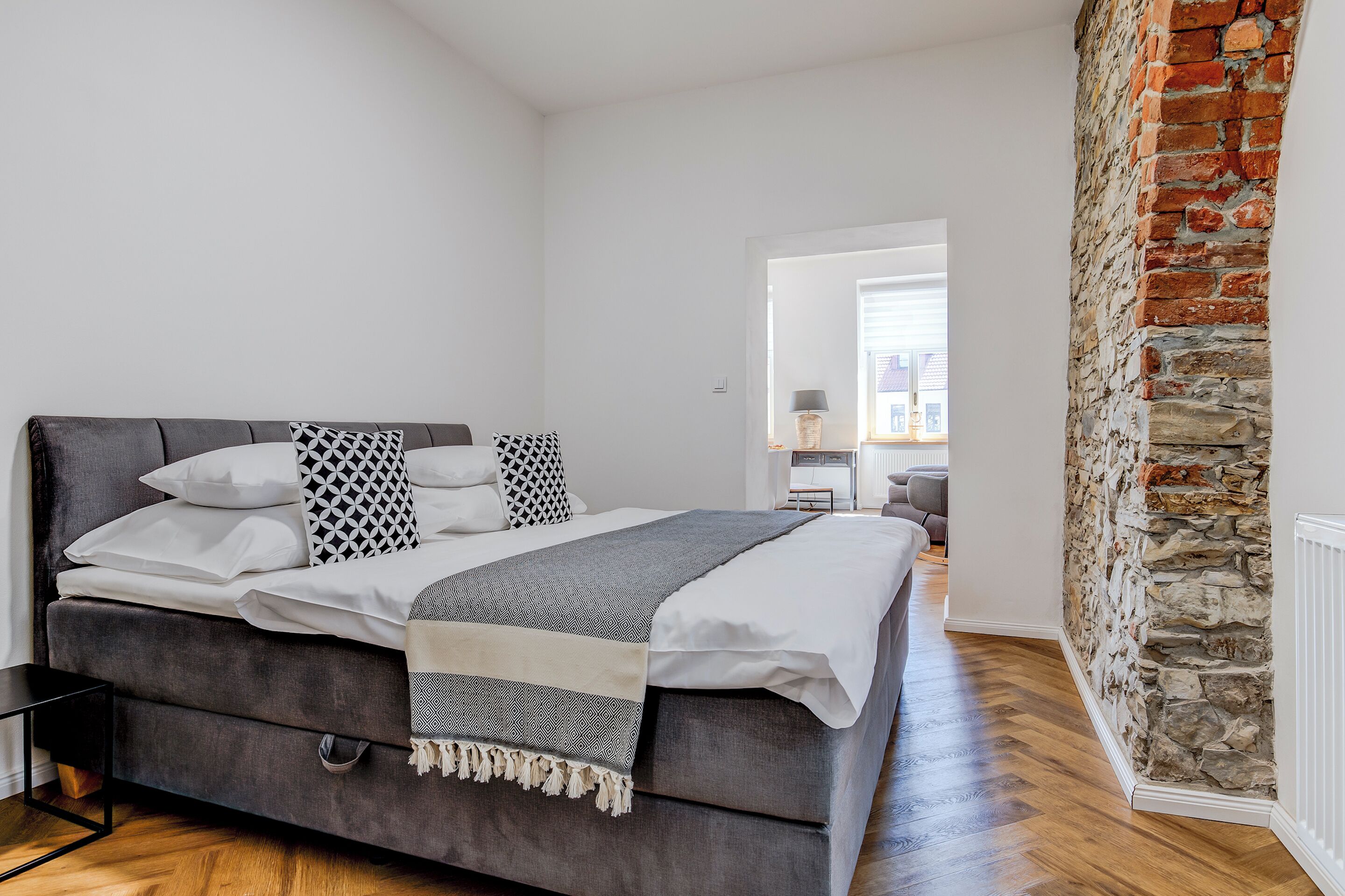 A modern bedroom with a grey upholstered bed, white linens, patterned pillows, and a grey throw blanket. A stone wall and wooden flooring are visible.