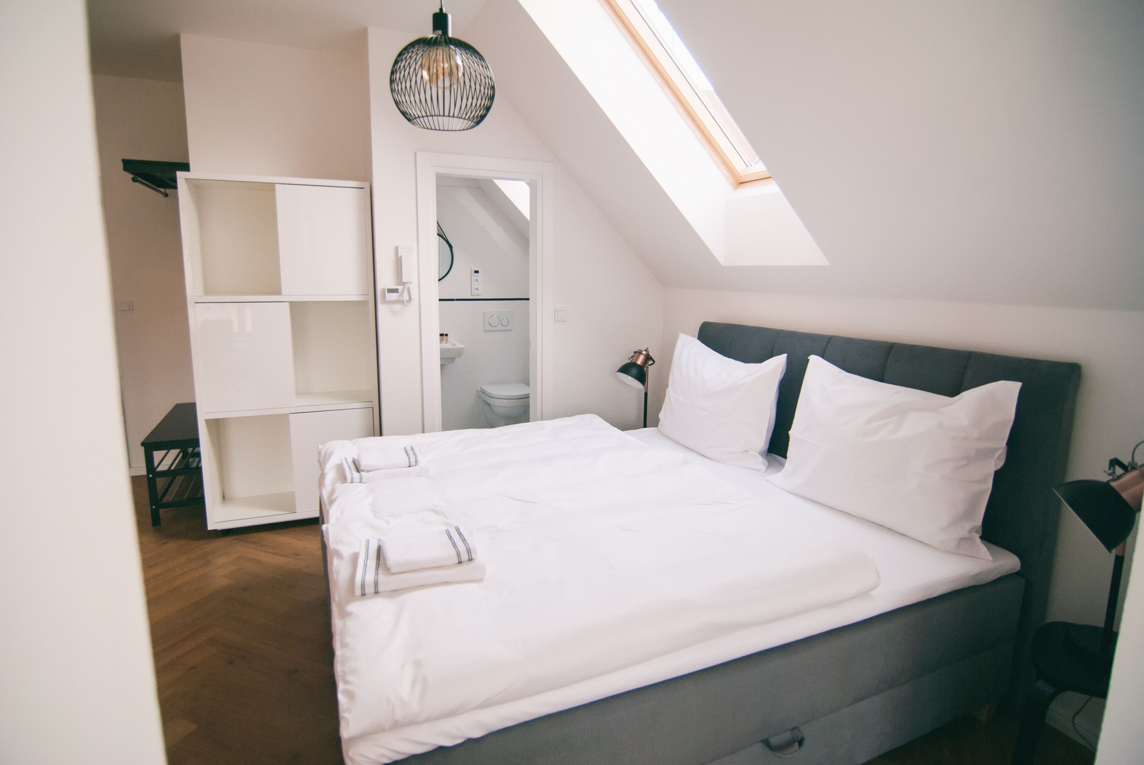 Ein Schlafzimmer mit Oberlicht, einem Bett mit weißer Bettwäsche, einem Nachttisch und einem Kleiderschrank.
