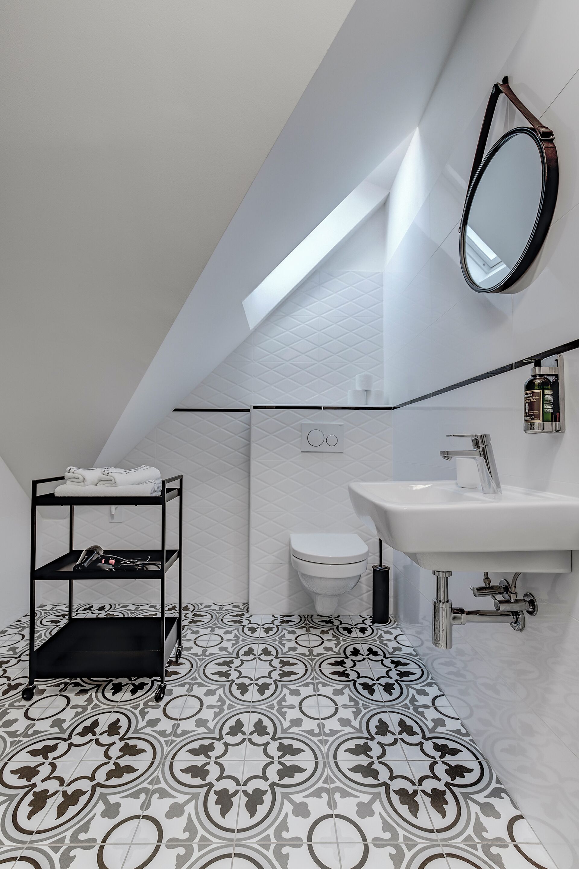 Un baño moderno con repisa metálica negra, lavamanos blanco y baldosas de suelo estampadas.
