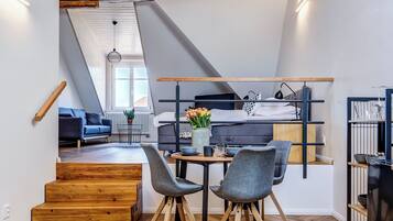 A modern loft-style room with exposed wooden beams, a herringbone wooden floor, a dining area with a table and chairs, a bunk bed, and a staircase leading to an upper level.