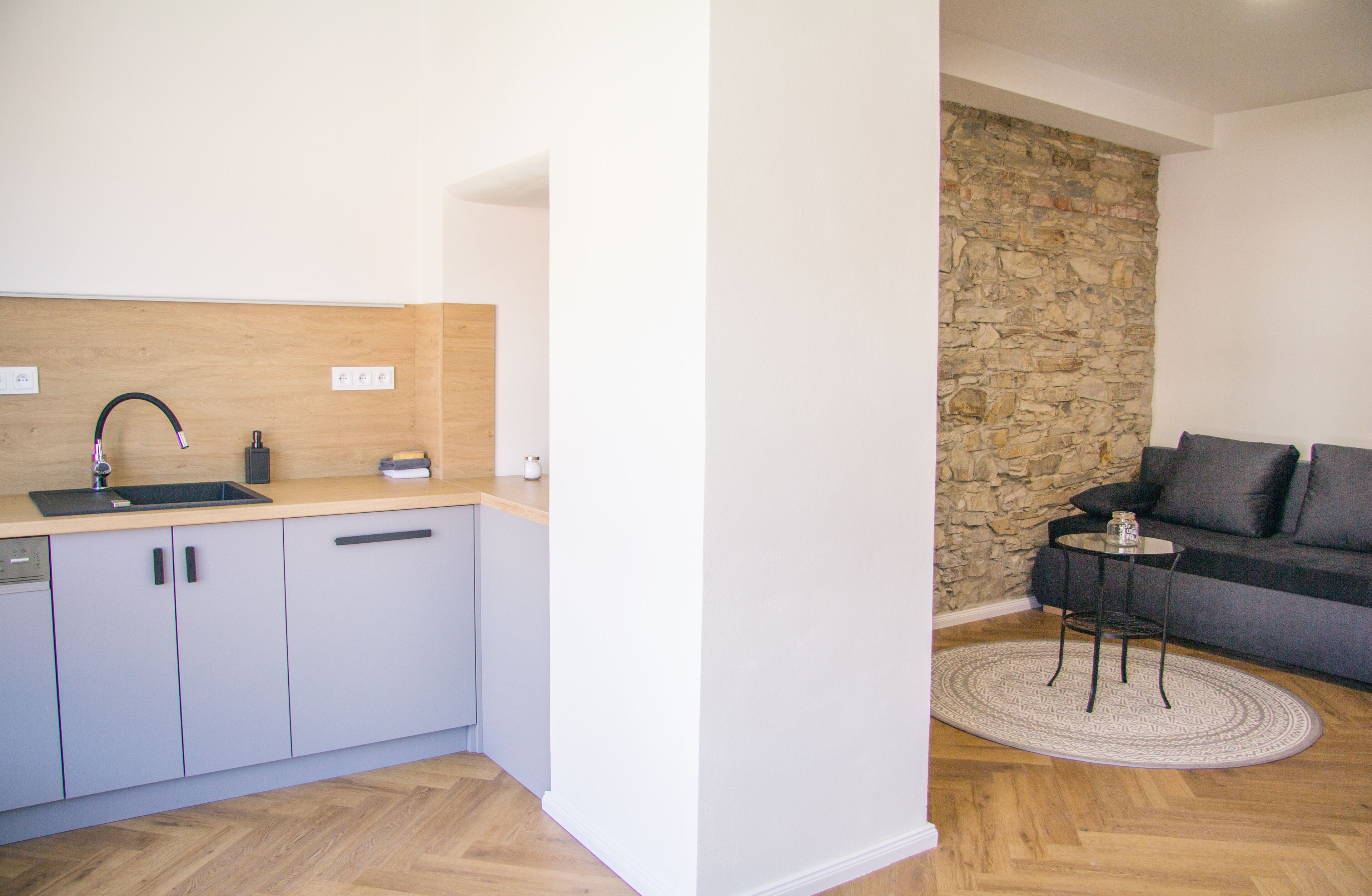 Una cocina moderna con encimera de madera y pared de piedra, junto a una sala de estar con sofá y una mesita redonda.