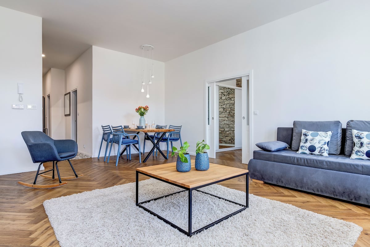 A modern living room with a wooden coffee table, a grey sofa with patterned cushions, and a dining area with a wooden table and blue chairs.
