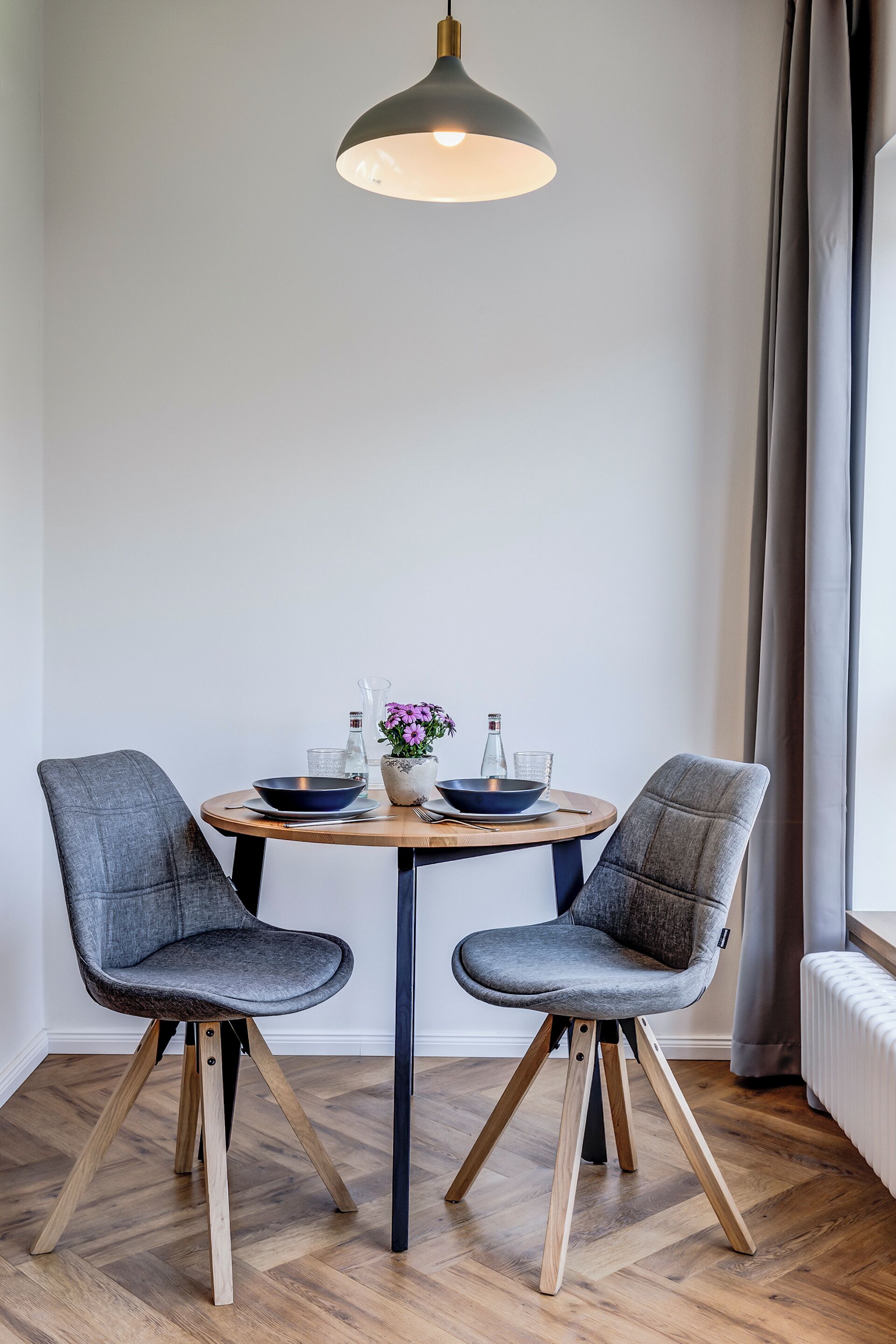 A dining area with a round table, two grey upholstered chairs, a pendant light, and a vase with flowers.