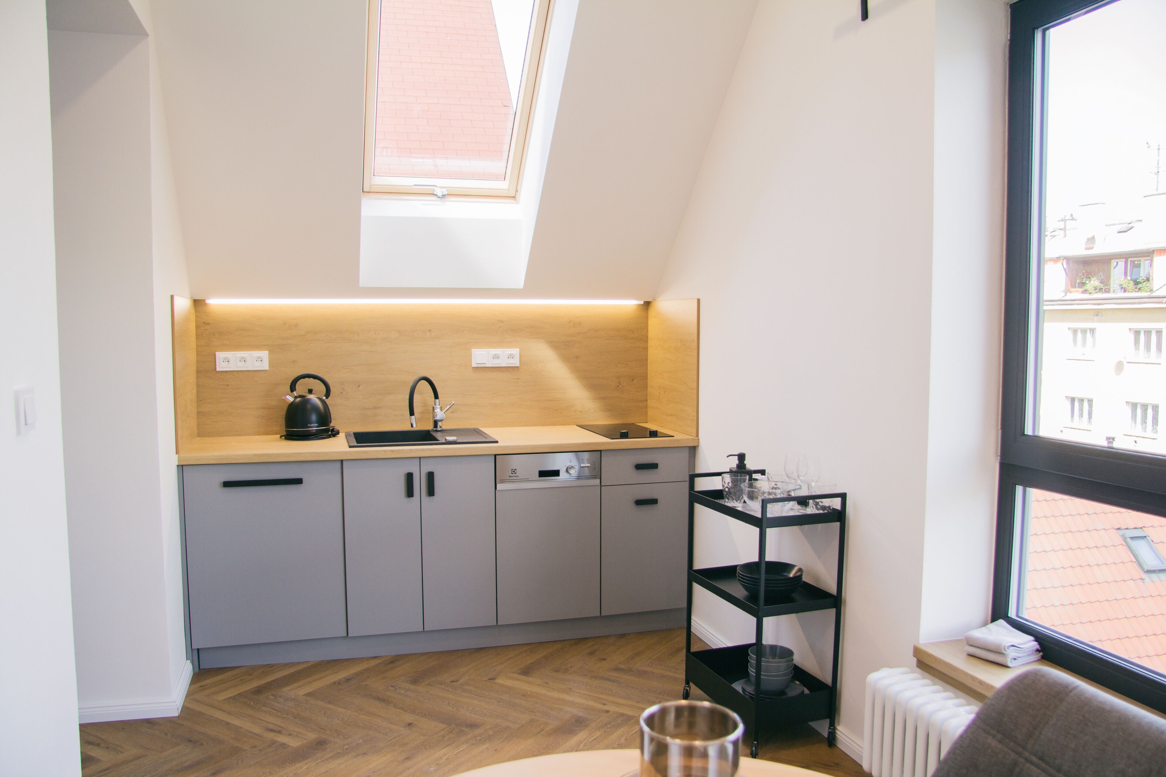 Une cuisine moderne avec des meubles gris, un plan de travail noir, une bouilloire et une fenêtre donnant sur des immeubles.