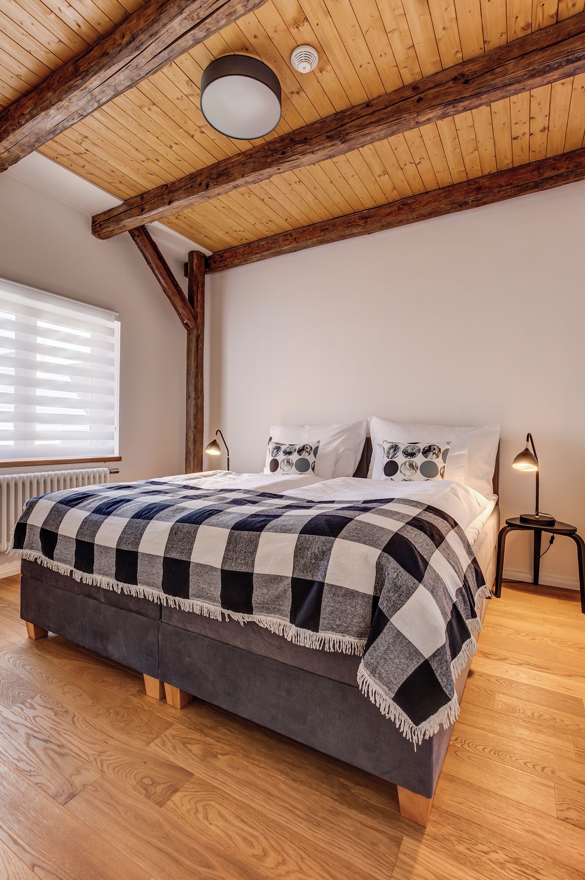 A bedroom with a wooden ceiling, a bed with a checkered blanket, a bedside lamp, and a window with blinds.