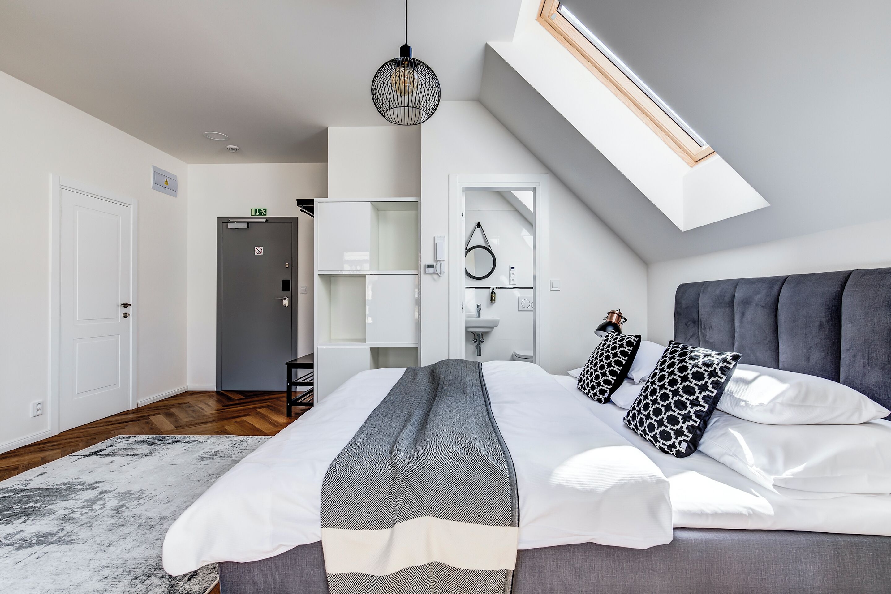 A modern bedroom with a large bed, a skylight, a wardrobe, and a bathroom visible through an open door.