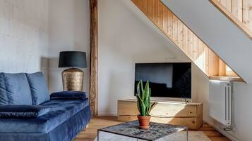 Et moderne loftværelse med synlige træbjælker, et stort vindue, en blå sofa, en gulvlampe og en potteplante.