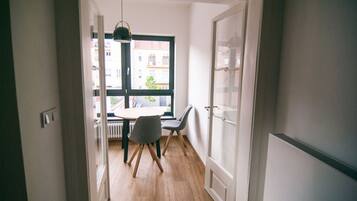 Un pasillo estrecho que conduce a un comedor con una mesa redonda y dos sillas, una ventana con vistas a edificios y una puerta con un panel de vidrio esmerilado.