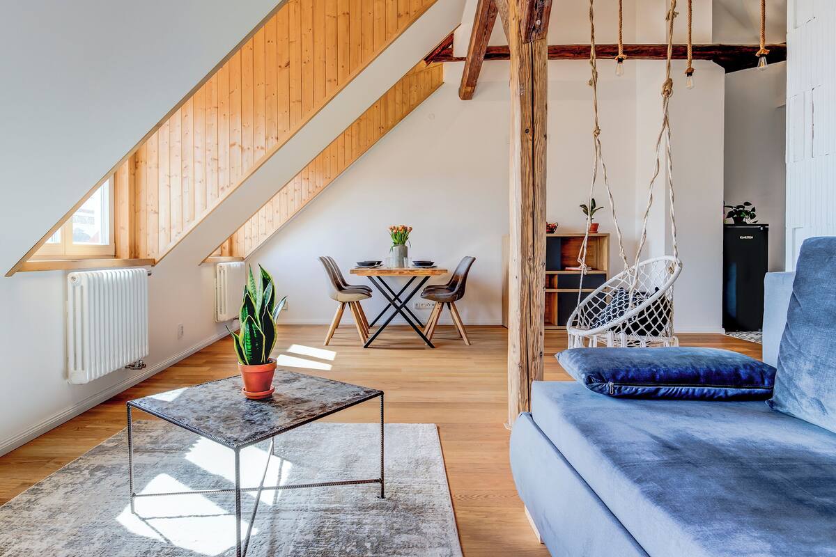 A loft-style room with a wooden ceiling, a dining area with a table and chairs, a hanging chair, and a blue sofa.