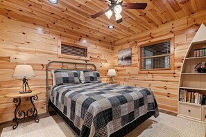 Quarto com painéis de madeira, uma cama, duas luminárias de cabeceira, um ventilador de teto e uma estante.