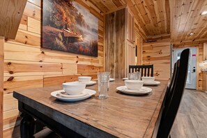 Interior de madeira com uma mesa de jantar posta para quatro pessoas, um quadro na parede e uma porta que dá para outro cômodo.