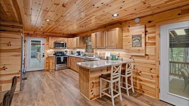 A wooden cabin interior with a kitchen featuring wooden cabinets, a central island with stools, a microwave, and a stove. - Beside Still Water 1 - 2 Bedrooms, 2 Baths, Sleeps 6 Home by RedAwning (Cosby)
