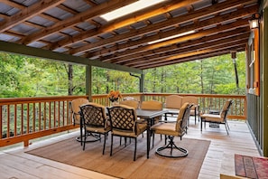 A covered outdoor deck with wooden railings, a table with chairs, and a view of a forested area. - Beside Still Water 1 - 2 Bedrooms, 2 Baths, Sleeps 6 Home by RedAwning (Cosby)