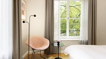 Una habitación con cama, una silla, una mesita con un florero y una ventana con vistas a árboles.