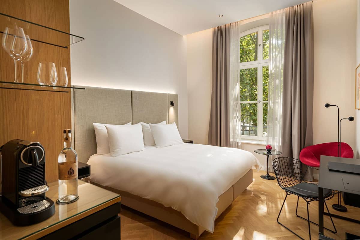 Ein modernes Hotelzimmer mit einem großen Bett, einem Schreibtisch mit Kaffeemaschine, einem roten Sessel und einem Fenster mit Vorhängen.