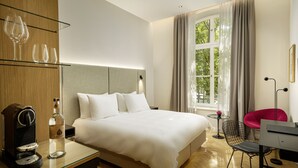 A modern hotel room with a large bed, a coffee machine, a glass table with wine glasses, a chair, and a view of greenery through the window. - ESPLANADE Saarbrücken (Saarbrücken)