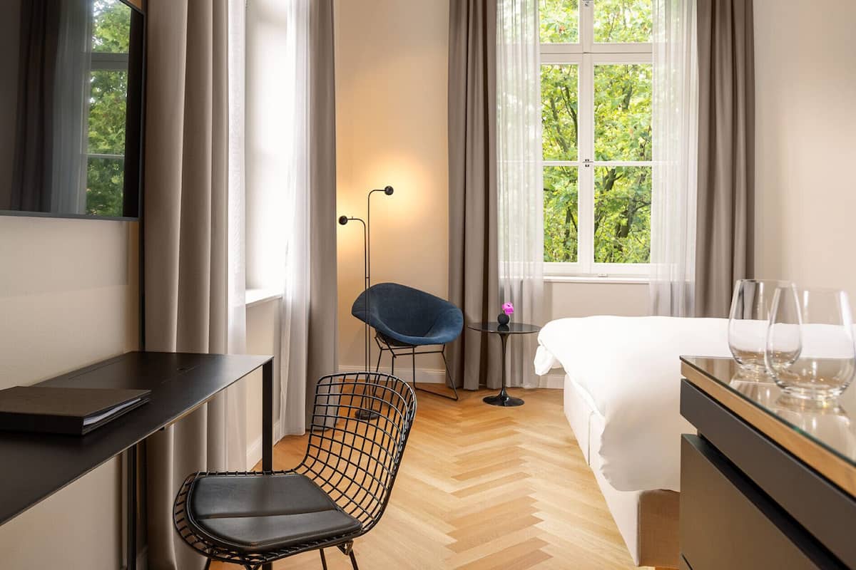 Ein Hotelzimmer mit Bett, Schreibtisch, Stuhl, kleinem Tisch mit Lampe und einem Fenster mit Vorhängen.