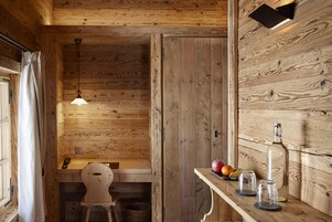 Interior de una cabaña de madera, con techo de madera, lámpara colgante, un banco de madera, una mesa con una botella, vasos y fruta.