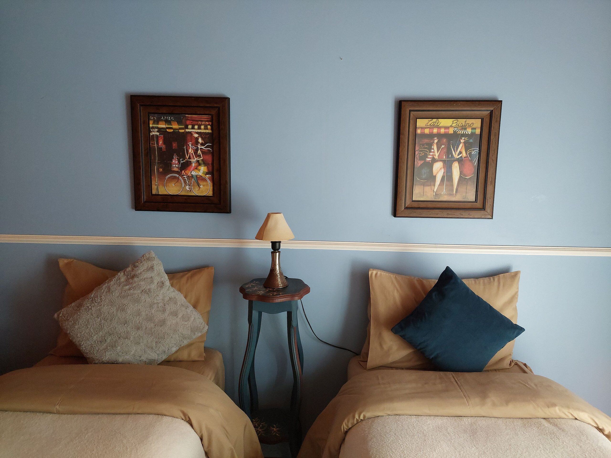 A room with two beds, each with a pillow and a lamp between them. There are two framed pictures on the wall above the beds.