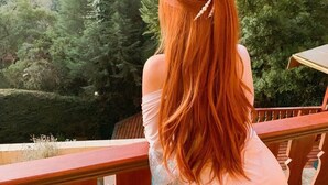 Una persona de cabello largo y rojizo sentada en un balcón con vista a un área boscosa y una casa al fondo.