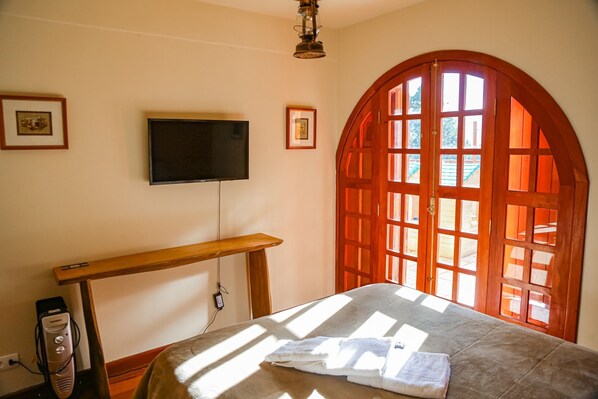 A room with a bed, a television mounted on the wall, and a large arched window with sunlight streaming in.