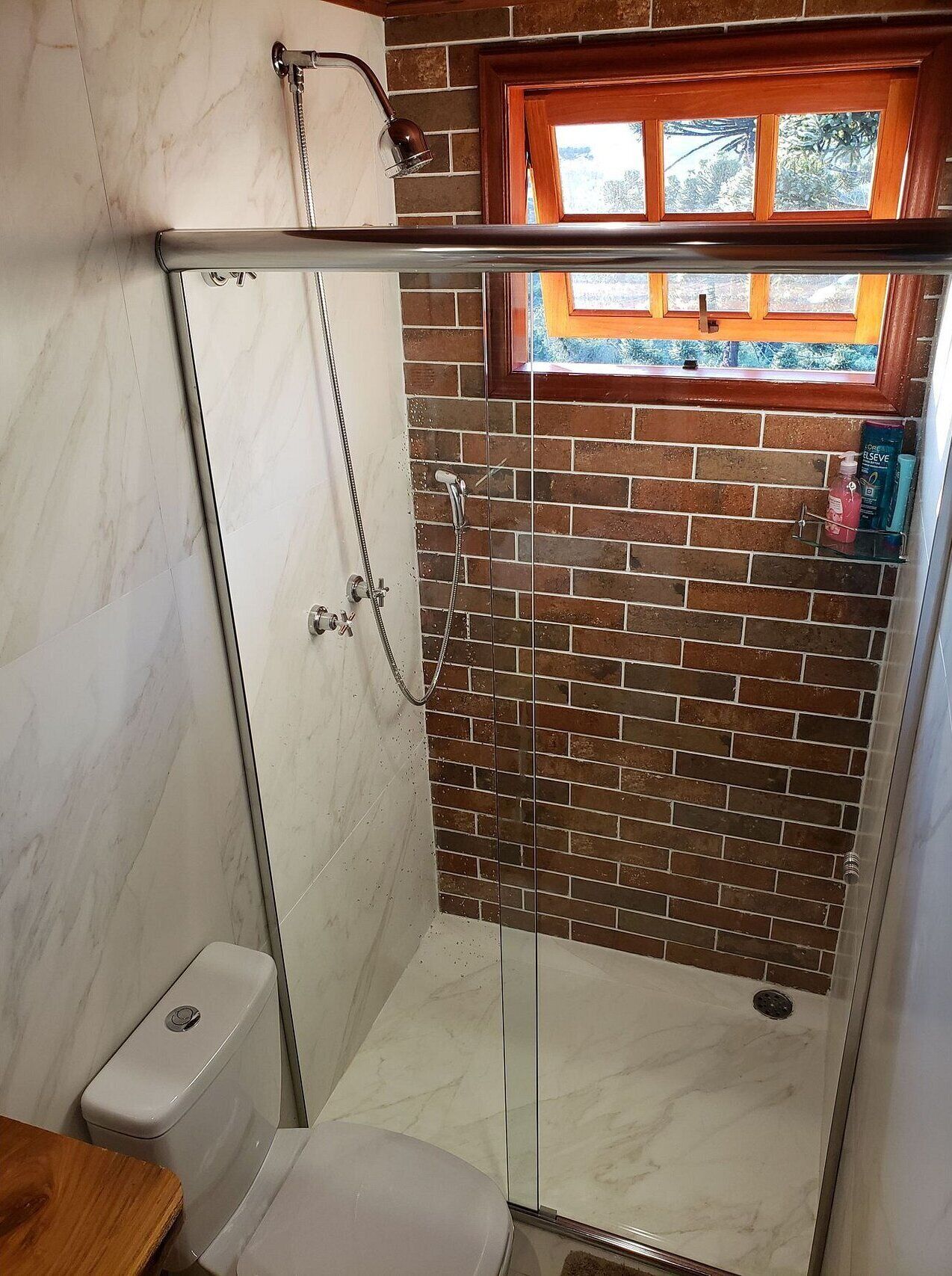 Een douche met glazen wanden, een betegelde bakstenen muur, een houten raam en een wit toilet.