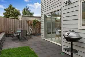 Un patio con mesa y sillas, una parrilla y un ventanal corredizo que da a un espacio interior.