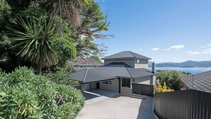 A modern two-story house with a covered patio, surrounded by lush greenery and a scenic view of the water. - Churchill House Riverside Retreat near Long Beach (Sandy Bay)