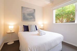 A neatly made bed with white bedding and a rolled towel, two bedside tables with lamps, a large abstract artwork on the wall, and a window with a view of trees. - Churchill House Riverside Retreat near Long Beach (Sandy Bay)