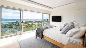 A bedroom with a large window, a bed with pillows, a flat-screen TV, and a view of a cityscape. - Churchill House Riverside Retreat near Long Beach (Sandy Bay)